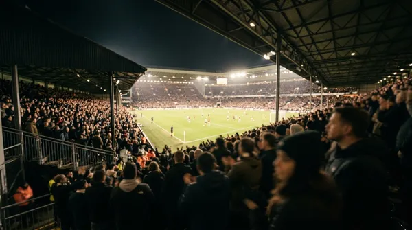 A floodlit football stadium at night seen from the stands, the crowd a warm blur